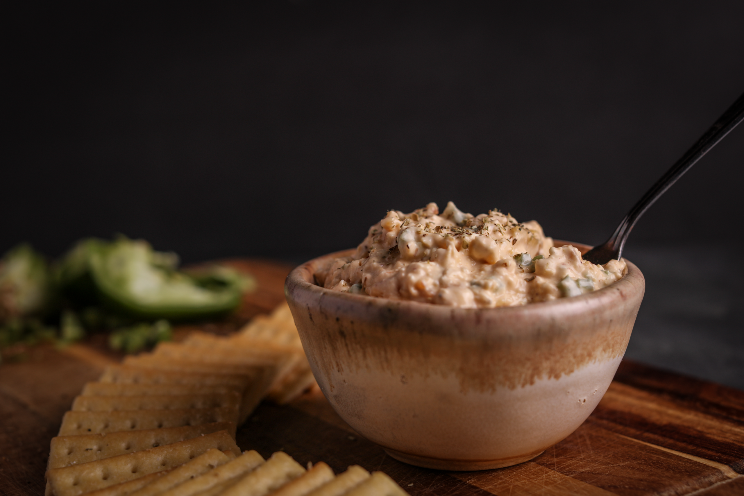 Bowl of Orange and Herb Cream Cheese dip in a tan and brown bowl on a cutting board with rectangular crackers fanned around the base and a diced bell pepper in the background