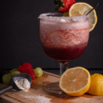 Front facing view of a sugar-rimmed goblet of pink and red ombre Blushing Berry Citrus Float Punch garnished with a lemon slice and skewer of grapes and a strawberry. Goblet is sitting on a cutting board surrounded by lemons, green grapes, and an ice cream scoop.