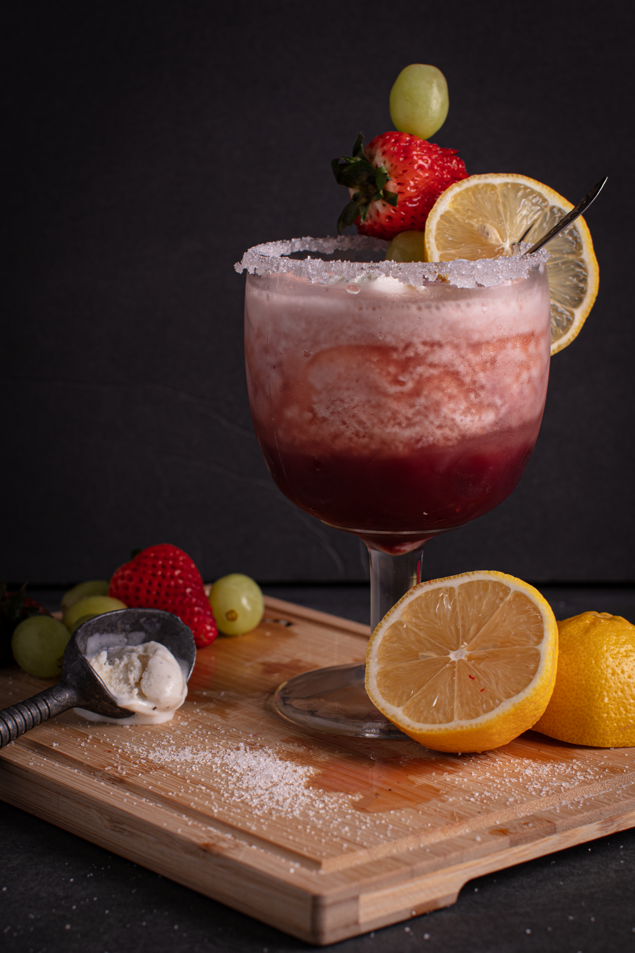 Front facing view of a sugar-rimmed goblet of pink and red ombre Blushing Berry Citrus Float Punch garnished with a lemon slice and skewer of grapes and a strawberry. Goblet is sitting on a cutting board surrounded by lemons, green grapes, and an ice cream scoop.
