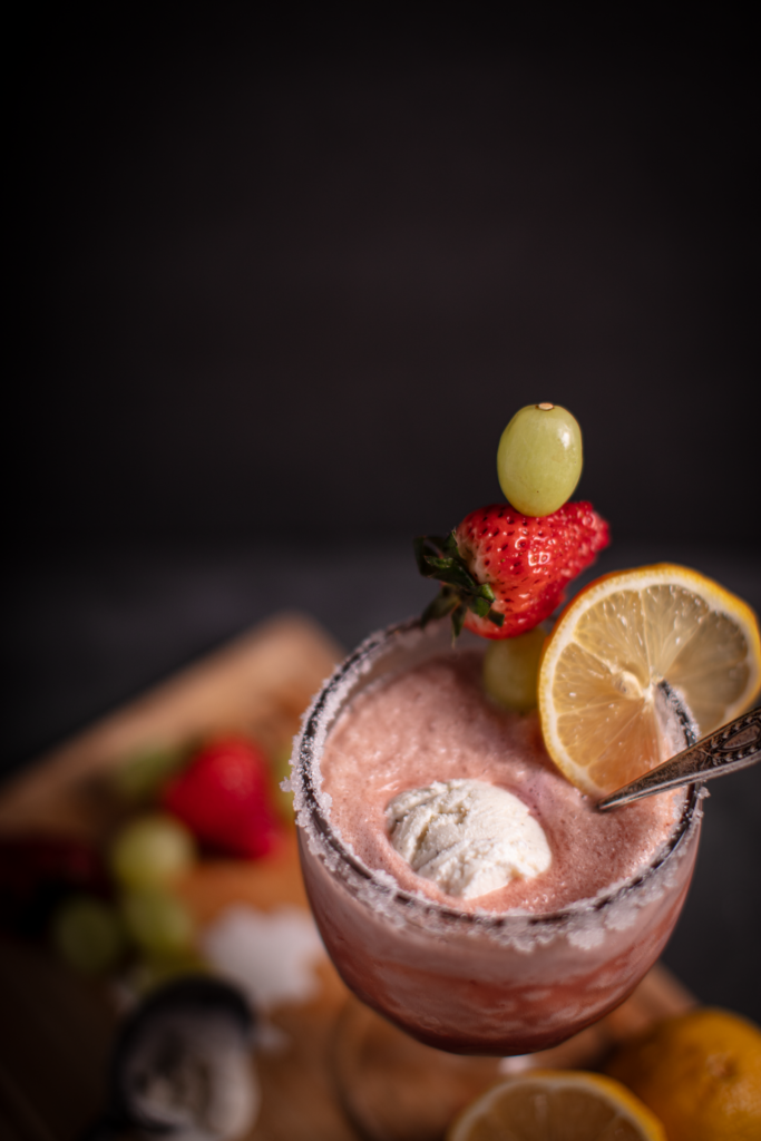 Closeup shot of Blushing Berry Citrus Float Punch in a sugar-rimmed goblet. The goblet is garnished with a slice of lemon and a skewer with green grapes and a strawberry.