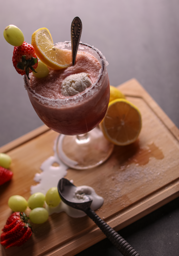 Goblet of frozen strawberry citrus drink with a scoop of vanilla ice cream. The rim of the goblet is sugared, and the drink is garnished with lemons, strawberries, and grapes. Grapes, lemons, strawberries, and an ice cream scoop surround the base of the goblet, which is standing atop a cutting board. The cutting board is sitting on a black background.