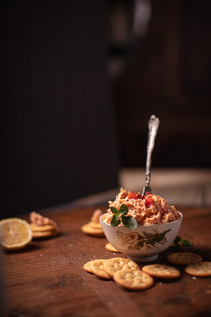 front view of a pimento cheese spread in a floral bowl with a spreader sticking out the top. crackers are nestled around the bottom of the bowl. all of this is on a wooden surface with a dark background