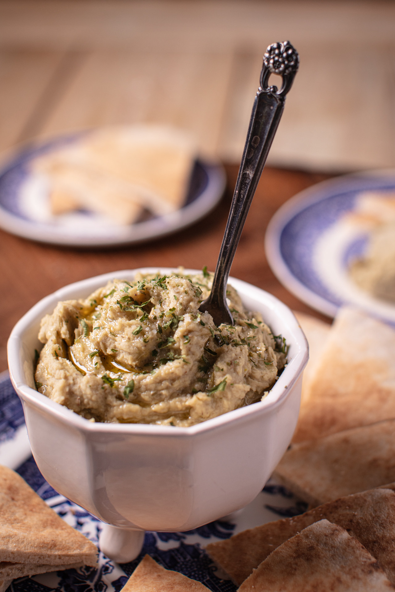butterbean and pistachio hummus in a white footed bowl with an antique silver spreader. The bowl is nestled about with pita triangles and sitting on a blue and white plate atop a wooden surface. Other blue and white appetizer plates with triangles of pita bread are blurred in the background