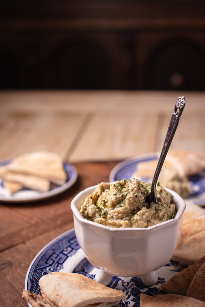 butterbean and pistachio hummus in a white footed bowl with a silver spreader sticking out. The bowl is on a blue and white plate with triangles of pita bread. Additional blue and white appetizer plates of pita bread can be see in the background atop the wooden surface.